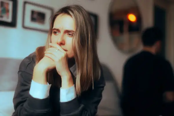 Kobieta zamyślona, z rękami przy twarzy, w tle niewyraźna postać. Może to symbolizować dystans emocjonalny w pracy, gdy czujemy się odizolowani.